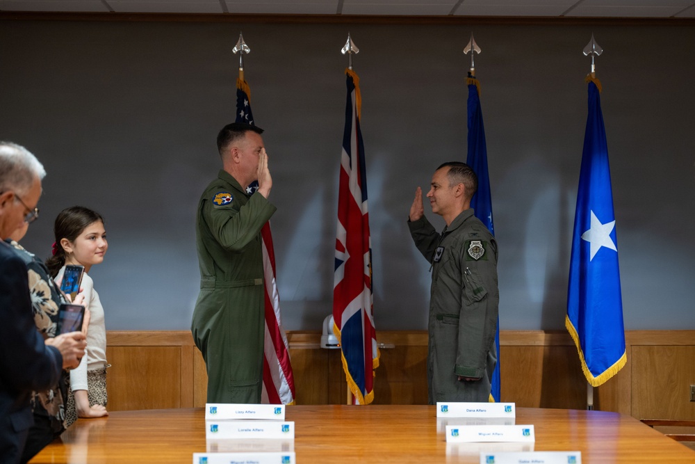 A military ceremony is taking place in a formal conference room, where two uniformed personnel in olive green flight suits are exchanging salutes. Name placards are visible on the table in front of them, suggesting a promotion or swearing-in ceremony. Family members, including a young girl holding a tablet, observe from the left side.