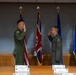A military ceremony is taking place in a formal conference room, where two uniformed personnel in olive green flight suits are exchanging salutes. Name placards are visible on the table in front of them, suggesting a promotion or swearing-in ceremony. Family members, including a young girl holding a tablet, observe from the left side.