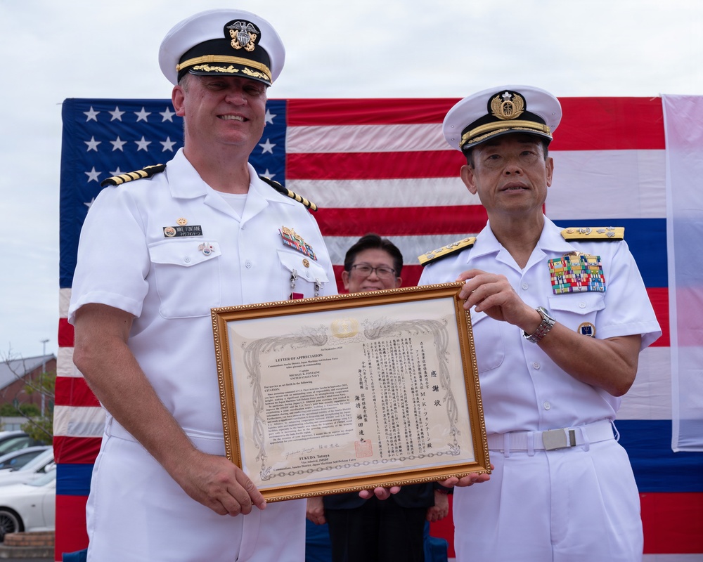 Two military officers in white dress uniforms — one from the United States Navy and one from the Japan Maritime Self-Defense Force (JMSDF) — are jointly holding a framed 'Letter of Appreciation' document during a formal ceremony. The certificate appears bilingual (English and Japanese) and is signed by FUKUDA Tatsuya, Commander, Sasebo District, Japan Maritime Self-Defense Force. An American flag is displayed prominently in the background.