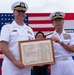Two military officers in white dress uniforms — one from the United States Navy and one from the Japan Maritime Self-Defense Force (JMSDF) — are jointly holding a framed 'Letter of Appreciation' document during a formal ceremony. The certificate appears bilingual (English and Japanese) and is signed by FUKUDA Tatsuya, Commander, Sasebo District, Japan Maritime Self-Defense Force. An American flag is displayed prominently in the background.