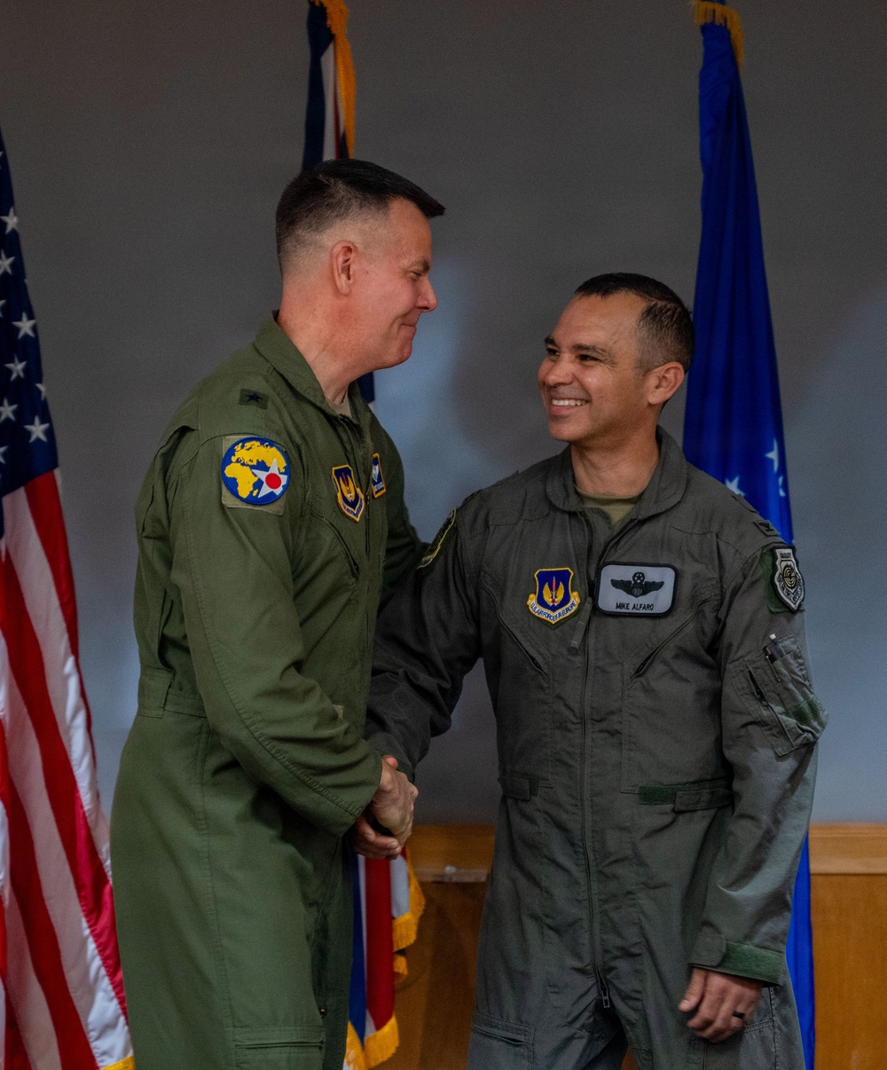 Two U.S. Air Force personnel in olive drab flight suits are shaking hands in what appears to be an official military ceremony. The individual on the right is identified by his name tag as 'Mike Alfaro' and wears a master pilot wing badge along with a USAF Reserve patch. The setting includes an American flag and a blue Air Force flag in the background.