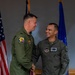 Two U.S. Air Force personnel in olive drab flight suits are shaking hands in what appears to be an official military ceremony. The individual on the right is identified by his name tag as 'Mike Alfaro' and wears a master pilot wing badge along with a USAF Reserve patch. The setting includes an American flag and a blue Air Force flag in the background.