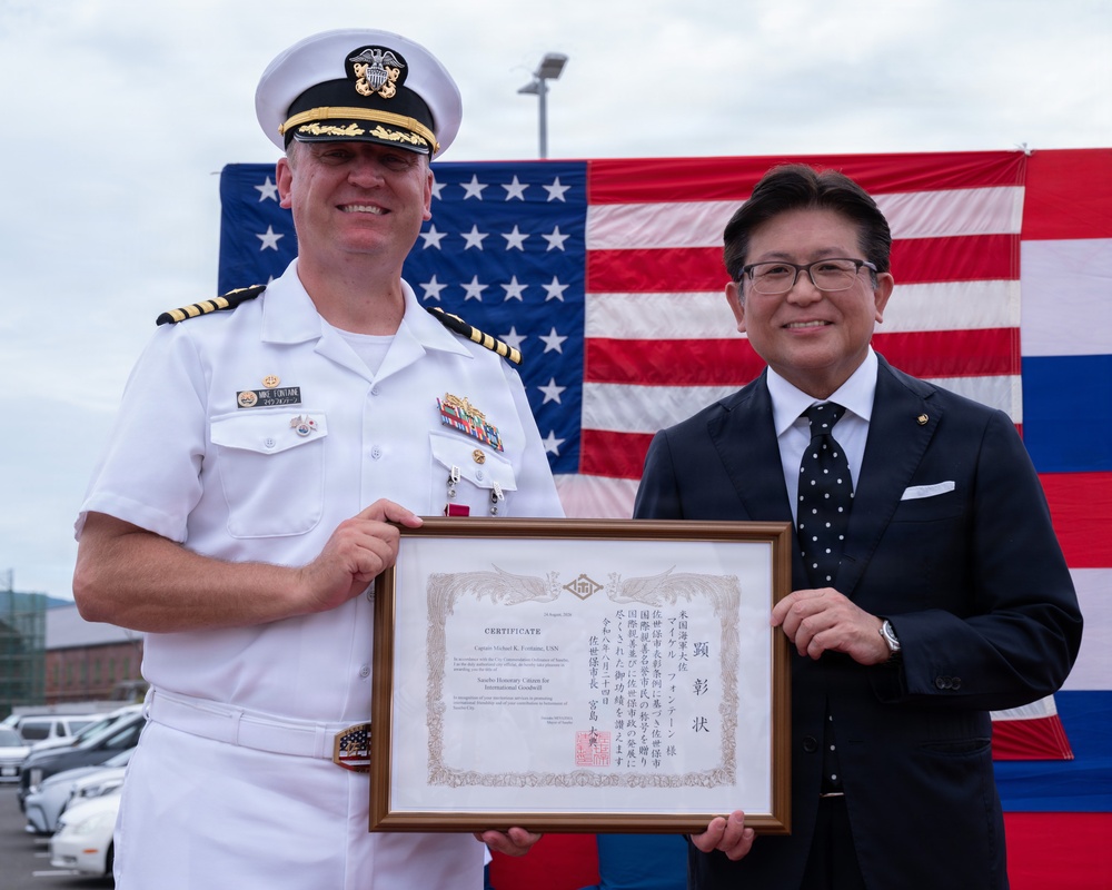 A U.S. Navy Captain in white dress uniform and a civilian in a dark suit jointly hold a framed certificate before an American flag backdrop. The certificate appears to be a 'Honorary Citizen for International Goodwill' award, with both English and Japanese text. The occasion appears to be a formal bilateral ceremony, likely in Japan.