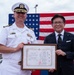 A U.S. Navy Captain in white dress uniform and a civilian in a dark suit jointly hold a framed certificate before an American flag backdrop. The certificate appears to be a 'Honorary Citizen for International Goodwill' award, with both English and Japanese text. The occasion appears to be a formal bilateral ceremony, likely in Japan.