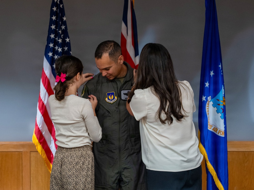 A U.S. Air Force service member in an olive green flight suit is having insignia or rank pinned onto his uniform by two females, likely family members, during a formal military ceremony. Three flags are visible in the background: the U.S. flag, a United Kingdom flag, and a U.S. Air Force flag, suggesting this may be taking place at a joint or overseas installation. The scene is consistent with a promotion or award ceremony.