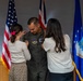 A U.S. Air Force service member in an olive green flight suit is having insignia or rank pinned onto his uniform by two females, likely family members, during a formal military ceremony. Three flags are visible in the background: the U.S. flag, a United Kingdom flag, and a U.S. Air Force flag, suggesting this may be taking place at a joint or overseas installation. The scene is consistent with a promotion or award ceremony.