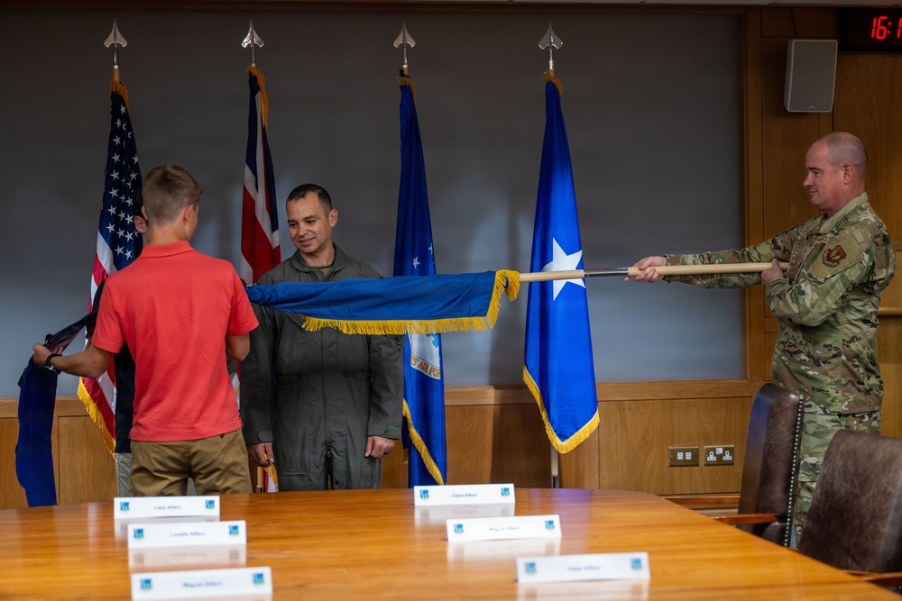 A military ceremony is taking place in a formal conference room, likely a promotion or award ceremony. Three individuals are handling a blue flag with gold fringe, which appears to be a US Air Force general officer flag featuring a single star. Name placards are visible on the conference table, suggesting family members are present for the occasion.