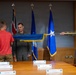 A military ceremony is taking place in a formal conference room, likely a promotion or award ceremony. Three individuals are handling a blue flag with gold fringe, which appears to be a US Air Force general officer flag featuring a single star. Name placards are visible on the conference table, suggesting family members are present for the occasion.