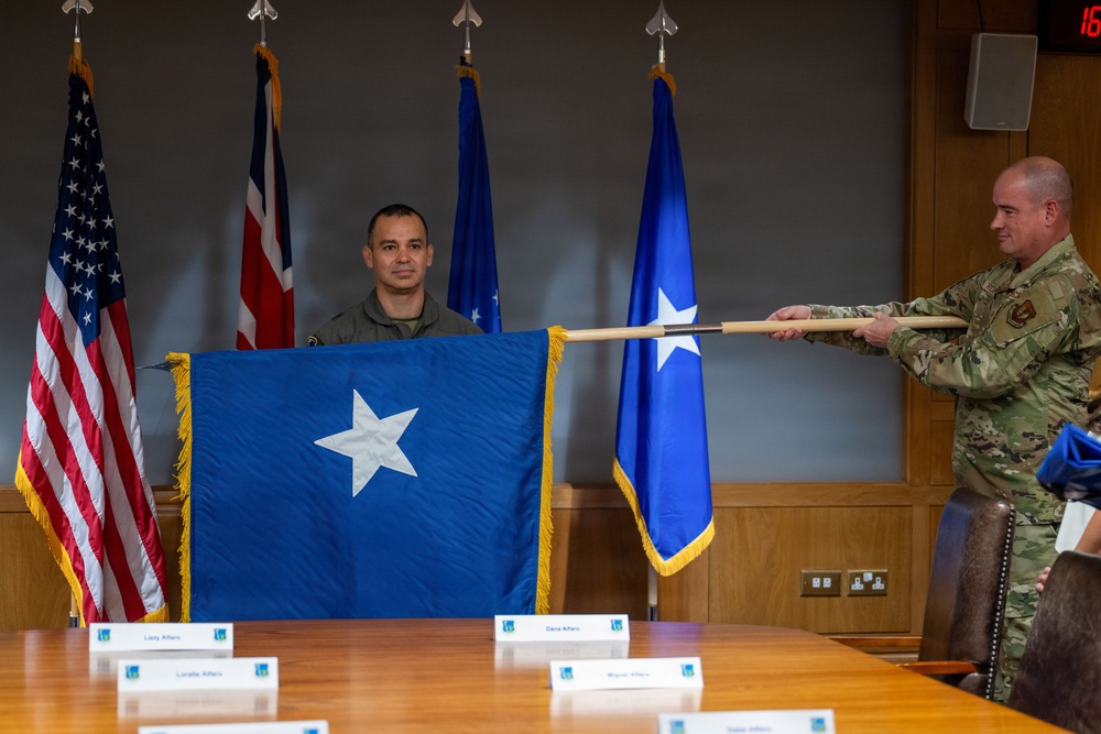 A military promotion ceremony is taking place in a formal conference room setting. Two uniformed personnel are displaying a blue one-star brigadier general flag with gold fringe, indicating a promotion to the rank of Brigadier General. Name placards are visible on the conference table, suggesting family members are present or expected for the ceremony.
