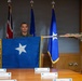 A military promotion ceremony is taking place in a formal conference room setting. Two uniformed personnel are displaying a blue one-star brigadier general flag with gold fringe, indicating a promotion to the rank of Brigadier General. Name placards are visible on the conference table, suggesting family members are present or expected for the ceremony.