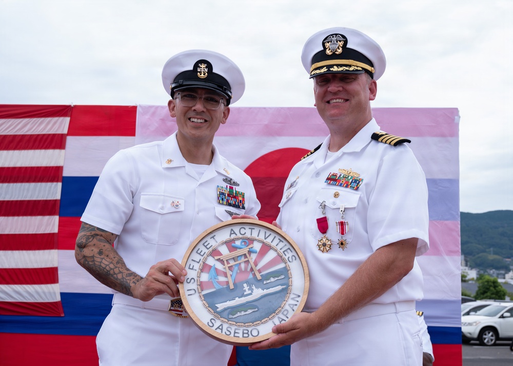 Two U.S. Navy personnel in white dress uniforms are holding a ceremonial plaque reading 'U.S. Fleet Activities Sasebo Japan' at what appears to be an official military ceremony. The backdrop features both the American flag and the Japanese flag, indicating a joint or bilateral military event. The occasion appears to be a change of command or award ceremony at Naval Station Sasebo, Japan.