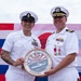 Two U.S. Navy personnel in white dress uniforms are holding a ceremonial plaque reading 'U.S. Fleet Activities Sasebo Japan' at what appears to be an official military ceremony. The backdrop features both the American flag and the Japanese flag, indicating a joint or bilateral military event. The occasion appears to be a change of command or award ceremony at Naval Station Sasebo, Japan.