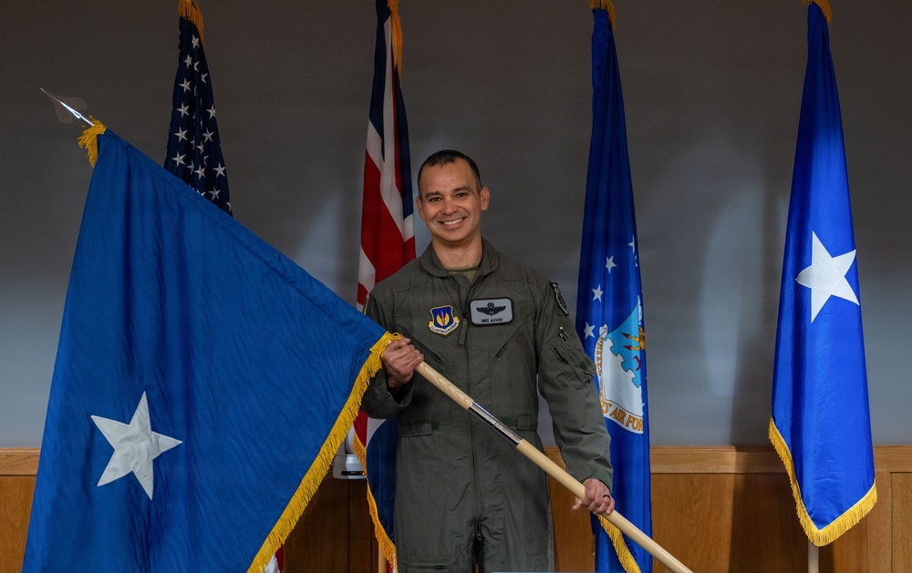 A U.S. Air Force officer in an olive green flight suit is posing while holding a blue one-star general officer flag with gold fringe. He is smiling and standing in front of multiple flags including the U.S. flag, a Union Jack (UK flag), and U.S. Air Force-related flags. The setting appears to be an official military ceremony or promotion event.