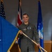 A U.S. Air Force officer in an olive green flight suit is posing while holding a blue one-star general officer flag with gold fringe. He is smiling and standing in front of multiple flags including the U.S. flag, a Union Jack (UK flag), and U.S. Air Force-related flags. The setting appears to be an official military ceremony or promotion event.