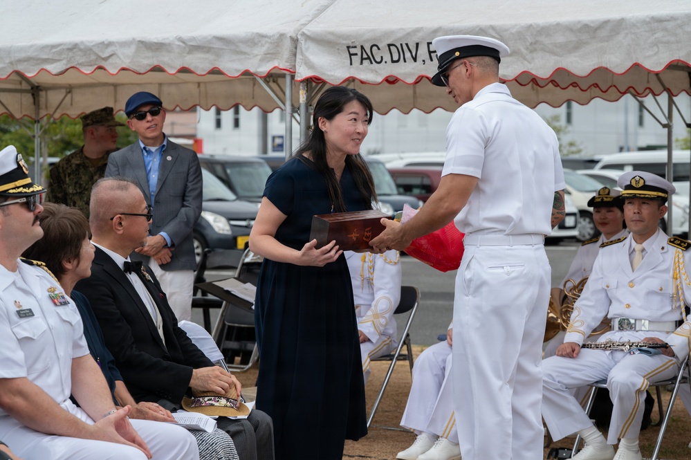 A formal military ceremony is taking place under a white tent, where a woman in a navy blue dress is receiving or exchanging a decorative wooden box with a naval officer in white dress uniform. Multiple military personnel in white uniforms, including what appear to be Japanese Maritime Self-Defense Force officers, are seated as observers. A banner partially reading 'FAC. DIV. F...' is visible in the background.