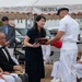 A formal military ceremony is taking place under a white tent, where a woman in a navy blue dress is receiving or exchanging a decorative wooden box with a naval officer in white dress uniform. Multiple military personnel in white uniforms, including what appear to be Japanese Maritime Self-Defense Force officers, are seated as observers. A banner partially reading 'FAC. DIV. F...' is visible in the background.