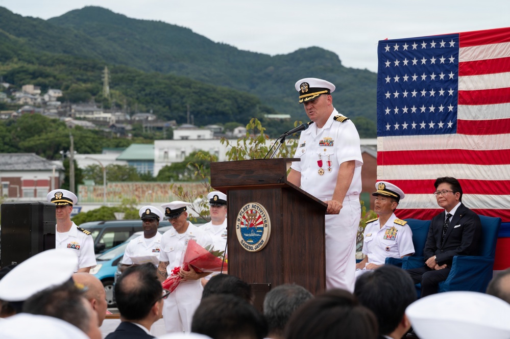 A U.S. Navy officer in white dress uniform delivers a speech at a podium bearing the seal of 'U.S. Fleet Activities Sasebo, Japan.' The ceremony takes place outdoors with an American flag displayed prominently in the background, alongside seated dignitaries including what appears to be a Japanese naval officer and a civilian official. Multiple Navy personnel in white uniforms stand in formation behind the speaker.