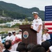 A U.S. Navy officer in white dress uniform delivers a speech at a podium bearing the seal of 'U.S. Fleet Activities Sasebo, Japan.' The ceremony takes place outdoors with an American flag displayed prominently in the background, alongside seated dignitaries including what appears to be a Japanese naval officer and a civilian official. Multiple Navy personnel in white uniforms stand in formation behind the speaker.