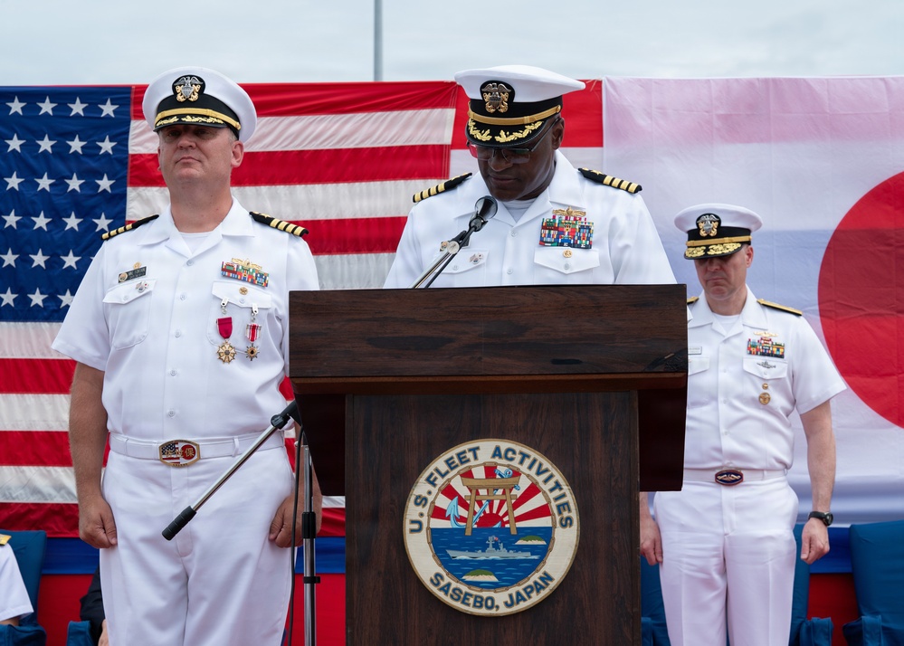 A formal U.S. Navy change of command or official ceremony is taking place at U.S. Fleet Activities Sasebo, Japan. Three naval officers in white dress uniforms are present, with one speaking at a podium bearing the U.S. Fleet Activities Sasebo, Japan seal. The American flag and Japanese flag are displayed prominently in the background.