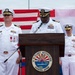 A formal U.S. Navy change of command or official ceremony is taking place at U.S. Fleet Activities Sasebo, Japan. Three naval officers in white dress uniforms are present, with one speaking at a podium bearing the U.S. Fleet Activities Sasebo, Japan seal. The American flag and Japanese flag are displayed prominently in the background.