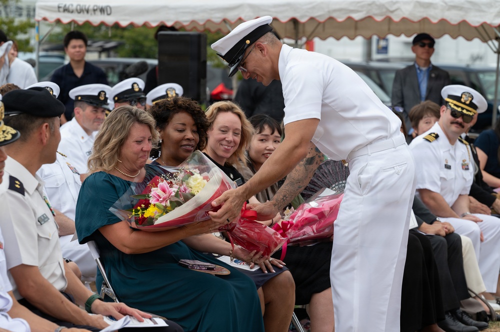 A U.S. Navy officer in dress white uniform is presenting a bouquet of flowers to a woman in a teal dress during what appears to be a formal military ceremony. The woman appears emotional, possibly tearful, while surrounded by other attendees including fellow Navy personnel in white uniforms and civilians. The event appears to be taking place outdoors under a tent, likely a change of command, retirement, or similar official military ceremony.