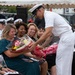 A U.S. Navy officer in dress white uniform is presenting a bouquet of flowers to a woman in a teal dress during what appears to be a formal military ceremony. The woman appears emotional, possibly tearful, while surrounded by other attendees including fellow Navy personnel in white uniforms and civilians. The event appears to be taking place outdoors under a tent, likely a change of command, retirement, or similar official military ceremony.