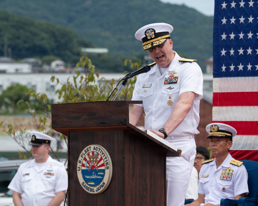 A senior U.S. Navy officer, wearing a white dress uniform with admiral-rank insignia and multiple service ribbons, is delivering a speech at a podium at U.S. Fleet Activities Sasebo, Japan. The event appears to be a formal military ceremony, with other naval personnel visible in the background alongside an American flag. The setting is an outdoor venue with green hills and base infrastructure visible behind the speakers.