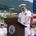 A senior U.S. Navy officer, wearing a white dress uniform with admiral-rank insignia and multiple service ribbons, is delivering a speech at a podium at U.S. Fleet Activities Sasebo, Japan. The event appears to be a formal military ceremony, with other naval personnel visible in the background alongside an American flag. The setting is an outdoor venue with green hills and base infrastructure visible behind the speakers.