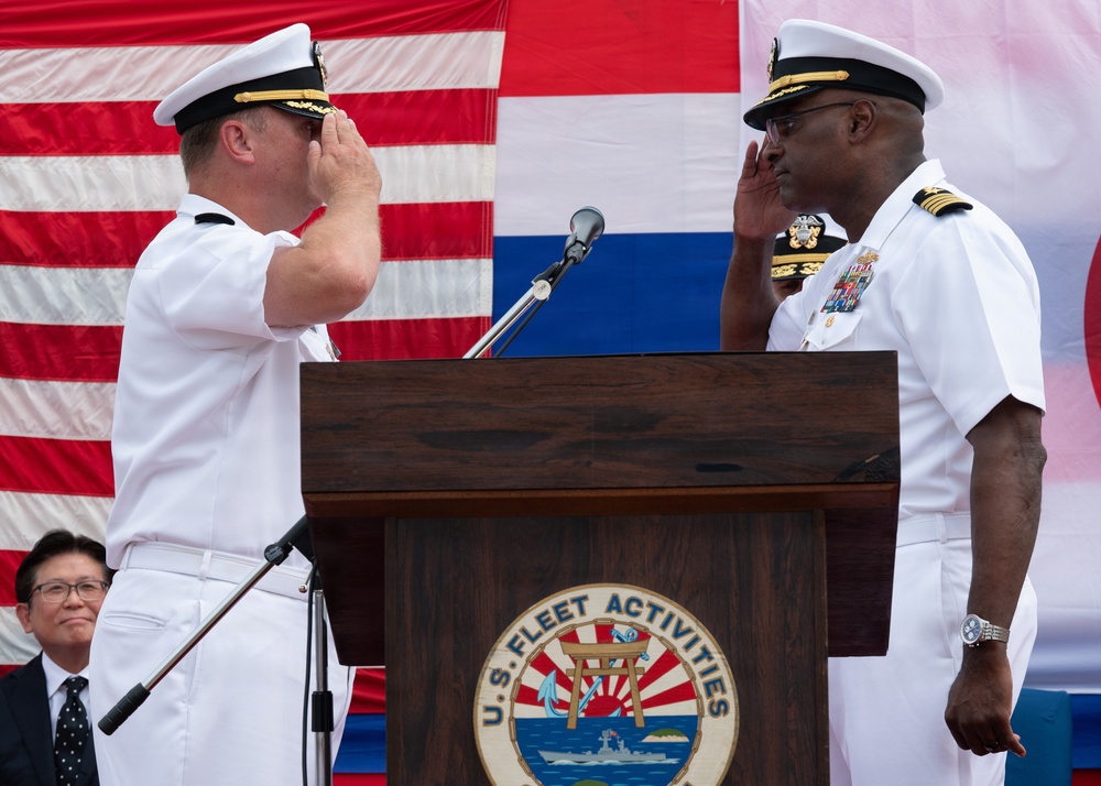 Two U.S. Navy officers in white dress uniforms are exchanging salutes at a podium bearing the U.S. Fleet Activities seal, likely during a change of command or official military ceremony. The event appears to take place outdoors with American and other flags visible in the background. A civilian observer is visible in the lower left corner.