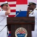 Two U.S. Navy officers in white dress uniforms are exchanging salutes at a podium bearing the U.S. Fleet Activities seal, likely during a change of command or official military ceremony. The event appears to take place outdoors with American and other flags visible in the background. A civilian observer is visible in the lower left corner.