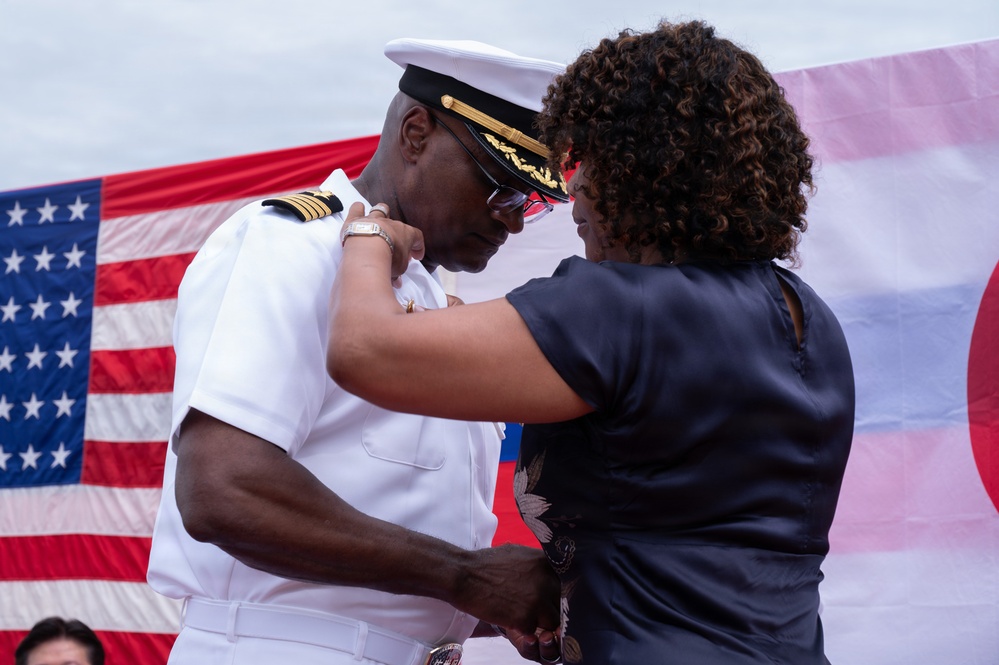 A U.S. Navy officer in white dress uniform is having insignia or decorations pinned onto his shoulder by a woman in a dark blouse. The ceremony appears to be a formal military promotion or commissioning event, taking place in front of an American flag. The interaction suggests a personal and ceremonial moment, likely involving a family member or colleague.