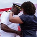 A U.S. Navy officer in white dress uniform is having insignia or decorations pinned onto his shoulder by a woman in a dark blouse. The ceremony appears to be a formal military promotion or commissioning event, taking place in front of an American flag. The interaction suggests a personal and ceremonial moment, likely involving a family member or colleague.