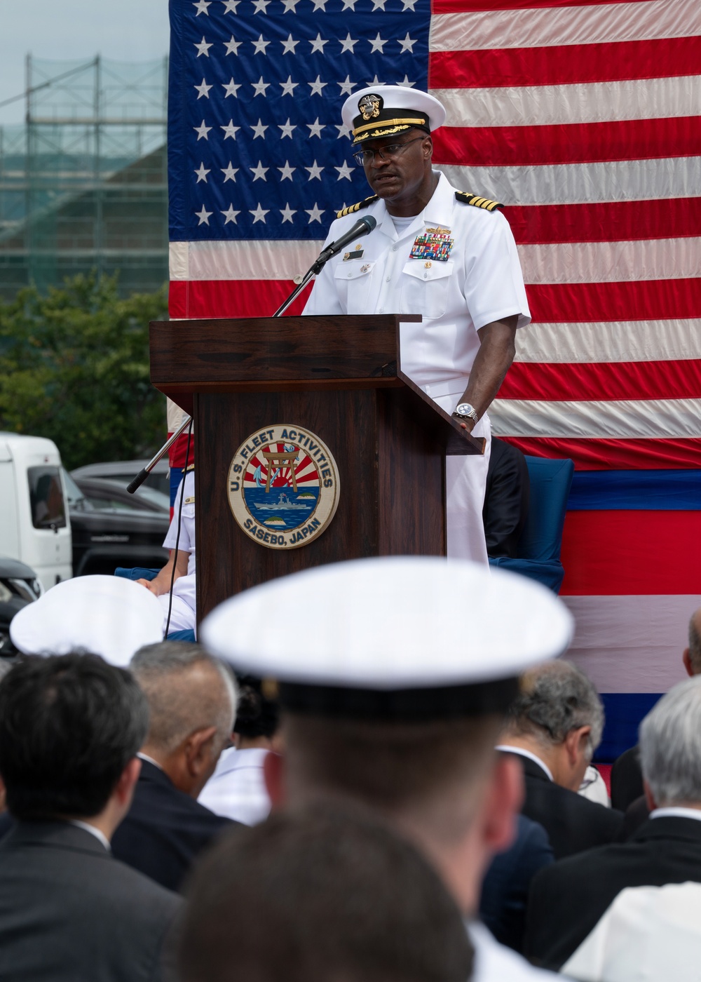 A U.S. Navy officer in white dress uniform is delivering a speech at a podium bearing the seal of U.S. Fleet Activities Sasebo, Japan. A large American flag serves as the backdrop, and an audience of military personnel and civilians is seated in the foreground. The event appears to be a formal military ceremony at the naval installation.