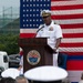 A U.S. Navy officer in white dress uniform is delivering a speech at a podium bearing the seal of U.S. Fleet Activities Sasebo, Japan. A large American flag serves as the backdrop, and an audience of military personnel and civilians is seated in the foreground. The event appears to be a formal military ceremony at the naval installation.