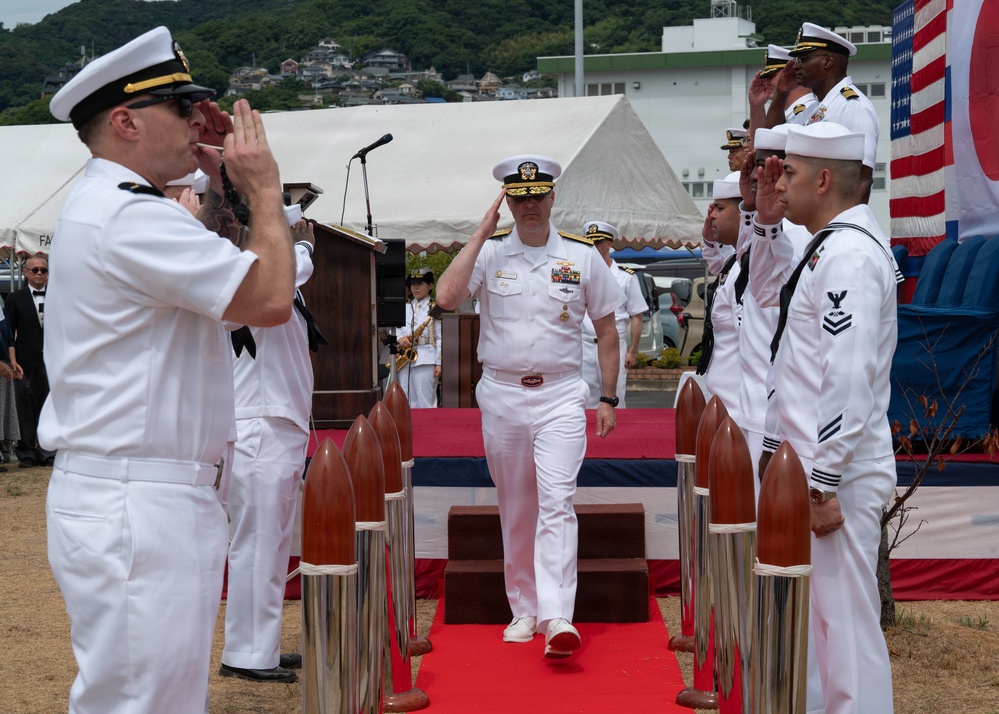 A formal U.S. Navy military ceremony is taking place outdoors, likely a change of command or similar official event. A senior officer in white dress uniform walks down a red carpet flanked by sideboys rendering salutes, with both the American and Japanese flags visible in the background. The setting appears to be a U.S. naval installation in Japan, suggested by the Japanese flag and surrounding landscape.