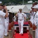 A formal U.S. Navy military ceremony is taking place outdoors, likely a change of command or similar official event. A senior officer in white dress uniform walks down a red carpet flanked by sideboys rendering salutes, with both the American and Japanese flags visible in the background. The setting appears to be a U.S. naval installation in Japan, suggested by the Japanese flag and surrounding landscape.