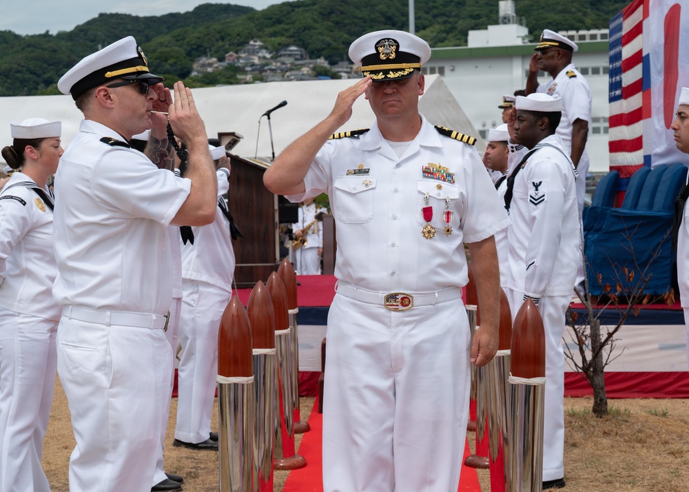 A formal U.S. Navy military ceremony is taking place outdoors, likely at a naval installation in Japan based on the hillside terrain and architecture visible in the background. A senior officer wearing a Captain's rank insignia and multiple service medals is rendering a salute, while a boatswain's mate appears to be piping (using a bosun's whistle) in a traditional sideboy honors ritual. Multiple sailors in dress white uniforms are standing in formation.