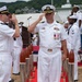 A formal U.S. Navy military ceremony is taking place outdoors, likely at a naval installation in Japan based on the hillside terrain and architecture visible in the background. A senior officer wearing a Captain's rank insignia and multiple service medals is rendering a salute, while a boatswain's mate appears to be piping (using a bosun's whistle) in a traditional sideboy honors ritual. Multiple sailors in dress white uniforms are standing in formation.