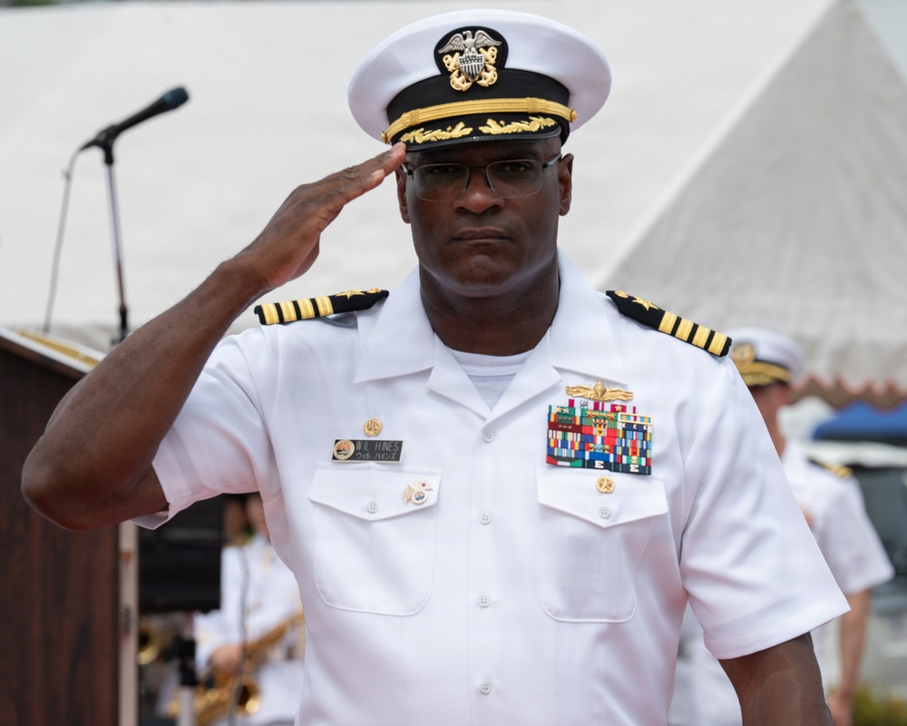 A senior U.S. Navy officer in white service dress uniform is rendering a formal salute at what appears to be an official military ceremony. The officer displays an extensive array of service ribbons and medals, along with gold warfare qualification devices, indicating significant military experience and achievement. The outdoor setting includes a tent canopy and other uniformed personnel visible in the background.