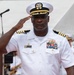 A senior U.S. Navy officer in white service dress uniform is rendering a formal salute at what appears to be an official military ceremony. The officer displays an extensive array of service ribbons and medals, along with gold warfare qualification devices, indicating significant military experience and achievement. The outdoor setting includes a tent canopy and other uniformed personnel visible in the background.