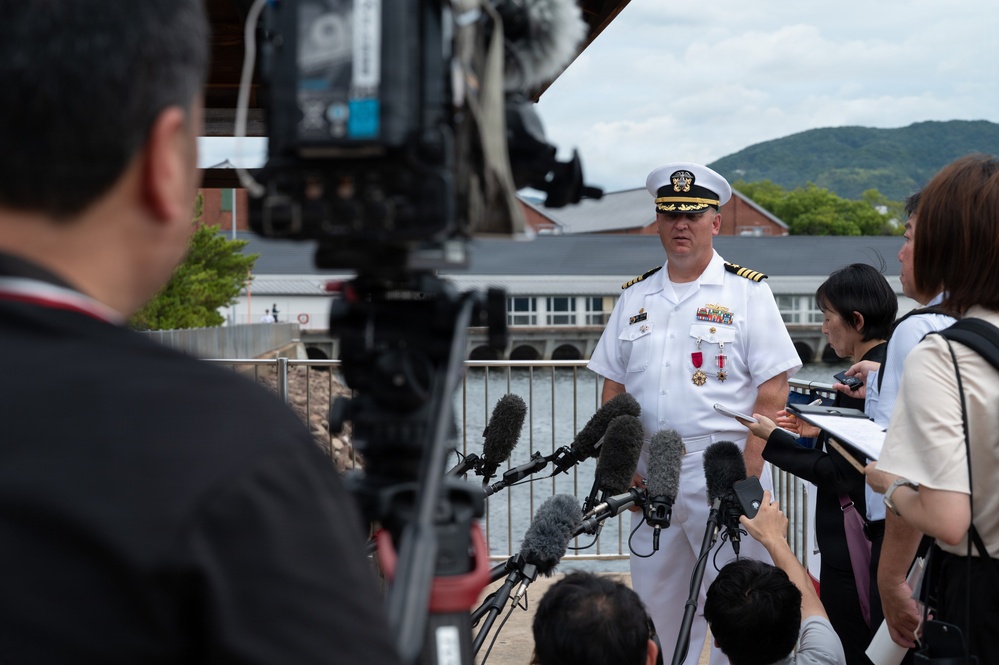 A U.S. Navy officer in white dress uniform is conducting a press conference or media briefing outdoors, surrounded by multiple journalists and camera operators. Several microphones are extended toward the officer, and a large broadcast camera is visible in the foreground. The setting appears to be near a waterfront in what looks like Japan, based on the architectural style and the apparent ethnicity of the journalists.
