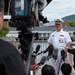 A U.S. Navy officer in white dress uniform is conducting a press conference or media briefing outdoors, surrounded by multiple journalists and camera operators. Several microphones are extended toward the officer, and a large broadcast camera is visible in the foreground. The setting appears to be near a waterfront in what looks like Japan, based on the architectural style and the apparent ethnicity of the journalists.