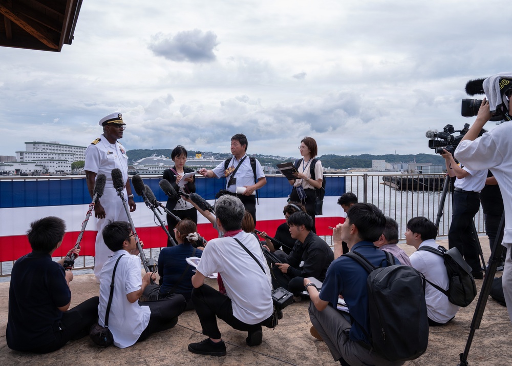 A U.S. Navy officer in white dress uniform is conducting a press conference at what appears to be a waterfront location, likely a Japanese port. A large group of journalists, predominantly Asian, are gathered around him with microphones, cameras, and recording equipment. A red, white, and blue barrier or banner is visible in the background, along with a harbor and cruise ship.