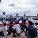 A U.S. Navy officer in white dress uniform is conducting a press conference at what appears to be a waterfront location, likely a Japanese port. A large group of journalists, predominantly Asian, are gathered around him with microphones, cameras, and recording equipment. A red, white, and blue barrier or banner is visible in the background, along with a harbor and cruise ship.