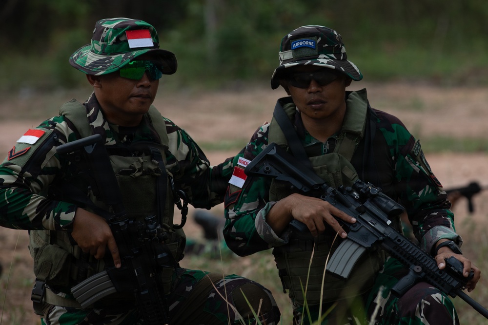 Two soldiers in Indonesian military (TNI) camouflage uniforms are seated in an outdoor field environment. Both wear tactical vests, sunglasses, and boonie hats — one displaying an Indonesian flag patch and the other an 'AIRBORNE' tab. The soldier on the right is holding an assault rifle, and both appear to be at rest during a training or operational exercise.
