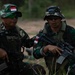 Two soldiers in Indonesian military (TNI) camouflage uniforms are seated in an outdoor field environment. Both wear tactical vests, sunglasses, and boonie hats — one displaying an Indonesian flag patch and the other an 'AIRBORNE' tab. The soldier on the right is holding an assault rifle, and both appear to be at rest during a training or operational exercise.