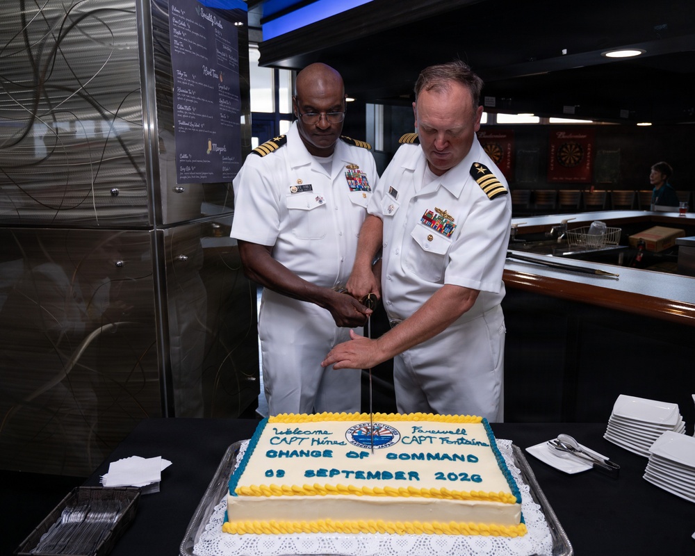 Two U.S. Navy Captains in white dress uniforms are cutting a ceremonial cake with a sword at a Change of Command ceremony. The cake identifies the incoming commander as CAPT Hines and the outgoing commander as CAPT Fontaine, dated 03 September 2026. The event appears to be held in a military club or dining facility.