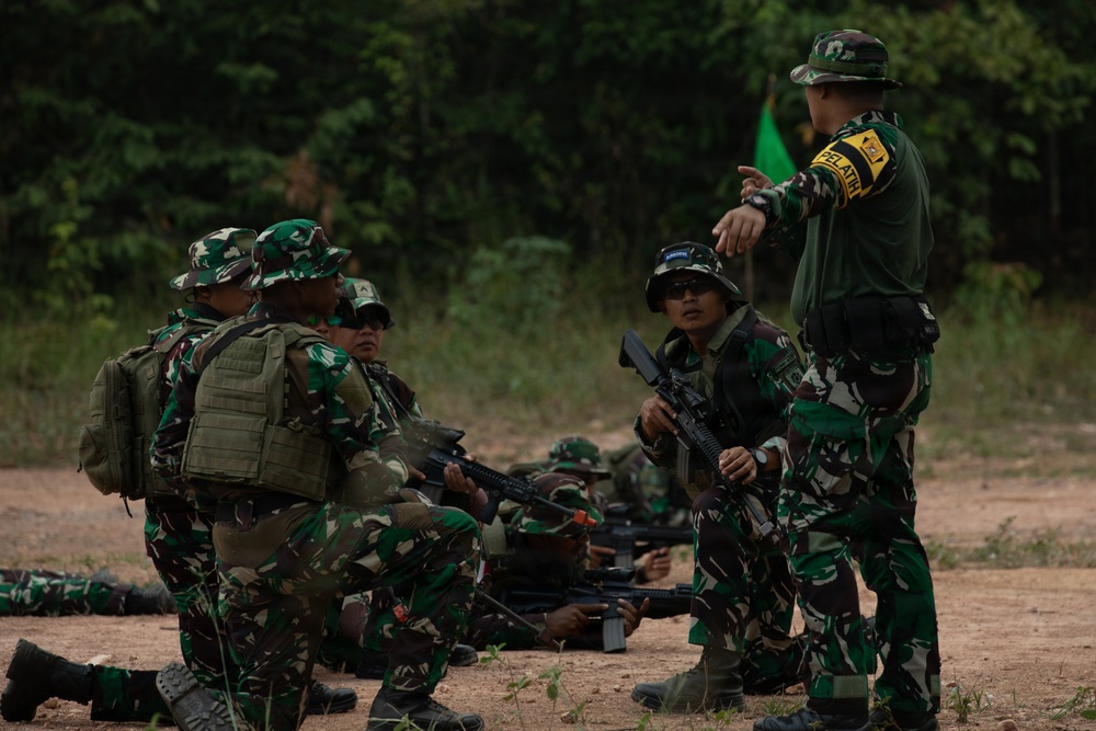 A military training exercise is underway in an outdoor forested area, with several soldiers in camouflage uniforms receiving instruction from a trainer. The standing instructor, identifiable by a yellow armband reading 'PELATIH' (Indonesian for 'trainer'), is directing soldiers who are positioned on the ground and in crouching stances. The soldiers are equipped with rifles and tactical gear, suggesting a field combat or tactical movement drill.