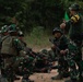 A military training exercise is underway in an outdoor forested area, with several soldiers in camouflage uniforms receiving instruction from a trainer. The standing instructor, identifiable by a yellow armband reading 'PELATIH' (Indonesian for 'trainer'), is directing soldiers who are positioned on the ground and in crouching stances. The soldiers are equipped with rifles and tactical gear, suggesting a field combat or tactical movement drill.