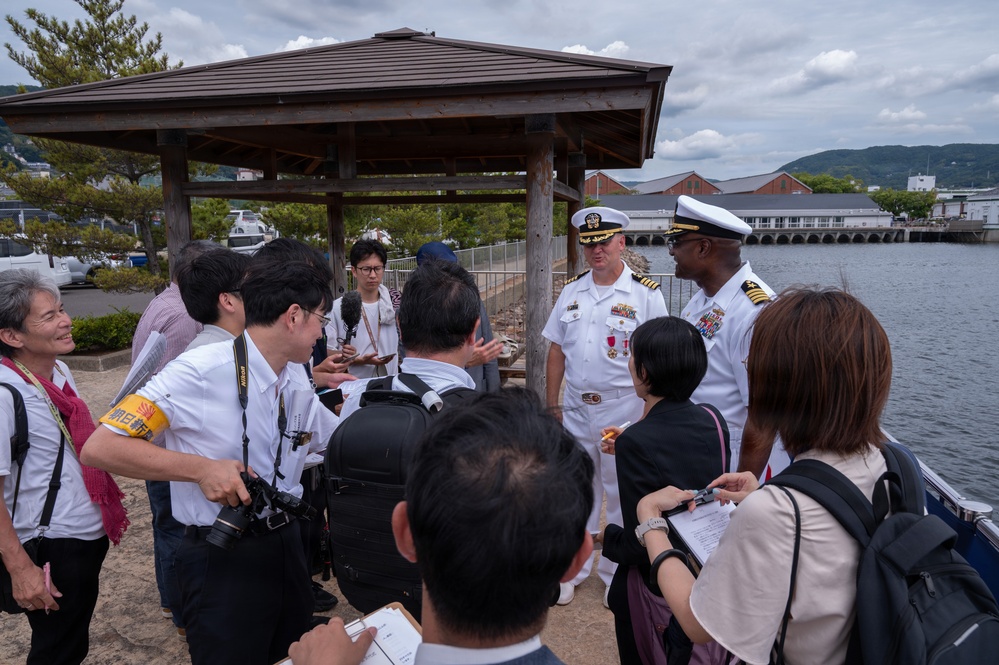 Two U.S. Navy officers in white dress uniforms are being interviewed or briefed by a group of Japanese civilians and journalists near a waterfront location, likely in Japan. The setting appears to be an outdoor area near a harbor or bay, with a wooden pavilion structure visible in the background. The interaction suggests a press engagement or community relations event.